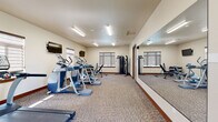 Located at 339 9th St in East Wenatchee, WA, this fitness center is a well-appointed, single-room exercise facility designed to support a comprehensive workout routine. The space is finished with neutral-toned carpeting and warm beige walls accented by rich wood baseboards, creating a clean and functional environment.

The room is equipped with a strong lineup of cardio machines, including multiple treadmills, ellipticals, and a stationary bike, all arranged to maximize use of the floor space. A dedicated strength training area features a multi-function cable machine and a dumbbell rack with a full set of free weights, complemented by an adjustable workout bench for versatile exercise options.

A full-length wall mirror runs along one side of the room, visually expanding the space and providing exercisers with proper form feedback during workouts. Multiple flat-screen TVs are mounted on the walls, offering entertainment during cardio sessions. Fluorescent linear lighting ensures the entire room is well-lit, while several windows fitted with zebra-style roller shades allow natural light to filter in. A hand sanitizer station near the entry reflects attention to cleanliness and user convenience. The overall layout is efficient and thoughtfully organized, making this fitness center a practical amenity for residents or guests.