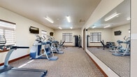 Located at 339 9th St in East Wenatchee, WA, this fitness center is a well-appointed, single-room exercise facility designed to support a comprehensive workout routine. The space is finished with neutral-toned carpeting and warm beige walls accented by rich wood baseboards, creating a clean and functional environment.

The room is equipped with a strong lineup of cardio machines, including multiple treadmills, ellipticals, and a stationary bike, all arranged to maximize use of the floor space. A dedicated strength training area features a multi-function cable machine and a dumbbell rack with a full set of free weights, complemented by an adjustable workout bench for versatile exercise options.

A full-length wall mirror runs along one side of the room, visually expanding the space and providing exercisers with proper form feedback during workouts. Multiple flat-screen TVs are mounted on the walls, offering entertainment during cardio sessions. Fluorescent linear lighting ensures the entire room is well-lit, while several windows fitted with zebra-style roller shades allow natural light to filter in. A hand sanitizer station near the entry reflects attention to cleanliness and user convenience. The overall layout is efficient and thoughtfully organized, making this fitness center a practical amenity for residents or guests.