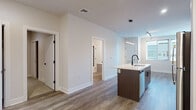 This contemporary single-floor residence showcases a clean, modern design with an open-concept layout that seamlessly connects the kitchen and living areas. The kitchen is a standout feature, equipped with stainless steel appliances including a refrigerator, electric range, and over-the-range microwave. A central island with a sleek white countertop and an integrated sink serves as both a functional workspace and a gathering point, accented by stylish pendant lighting above. Cabinetry combines crisp white upper cabinets with dark wood-toned lower cabinets, complemented by a subtle tile backsplash. Wood-look luxury vinyl plank flooring flows throughout the main living areas, adding warmth and continuity to the space. The bedroom offers plush carpeting, natural light through a set of windows with blinds, and a calm, neutral palette. The bathroom features a tub-shower combination enclosed by sliding glass doors with matte black hardware, paired with a white countertop vanity and wood-toned cabinetry. Throughout the residence, matte black hardware and door fixtures provide a cohesive, contemporary accent. The overall ambiance is bright, clean, and move-in ready, reflecting a thoughtfully designed modern living space.