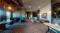 This elegantly designed single-floor communal space blends modern sophistication with warm, inviting details across several distinct functional zones. The herringbone-patterned carpet flows throughout, tying together the various areas with a cohesive visual rhythm. Exposed dark wood beams accent the ceiling, adding architectural character and a grounded, refined atmosphere.

The lounge area serves as the social heart of the space, anchored by a sleek fireplace framed by textured wallcovering and flanked by teal upholstered accent chairs. Nearby, a tufted leather sofa pairs with vibrant mustard yellow velvet chairs, centered around a low coffee table beneath a striking world map artwork mounted on a dimensional, geometric-textured white wall. Built-in dark cabinetry with diamond-pattern open shelving provides both display and storage, adding depth and visual interest.

A dedicated meeting or dining zone features a long, dark wood communal table surrounded by rust-toned swivel chairs, illuminated dramatically by a series of sculptural wood-framed pendant lights. Floor-to-ceiling windows flood this area with natural light and offer views of surrounding greenery. A wall-mounted flat-screen display makes the space well-suited for presentations or collaborative work.

A cozy alcove with a velvet banquette and bistro table offers a more intimate setting, while the overall design palette of deep charcoals, warm woods, and jewel-toned accents creates a polished, upscale ambiance throughout.