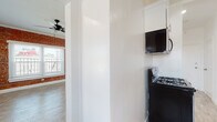 This single-floor space combines classic character with contemporary finishes throughout. The living area serves as the centerpiece of the unit, showcasing a striking full wall of exposed red brick that adds warmth and industrial charm. Multiple windows with white blinds flood the room with natural light, while crown molding, recessed lighting, and a ceiling fan contribute to a polished yet relaxed ambiance. Light wood-tone flooring runs seamlessly throughout the space, unifying each area.

The galley-style kitchen is efficiently designed with crisp white shaker cabinets, dark countertops, and brushed nickel hardware. Black appliances — including a gas range with overhead microwave and a full-size refrigerator — provide a bold contrast to the bright cabinetry. A single-basin stainless steel sink with a modern faucet completes the functional layout.

The bathroom features bold marble-look tile surround at the tub/shower, paired with light wood-tone flooring and a white vanity, creating a clean and elegant feel. A frosted window allows natural light while maintaining privacy.

An additional compact room with white walls and light wood-tone flooring offers flexible use as a bedroom, office, or storage space. Overall, the unit presents a cohesive blend of exposed brick character and updated modern finishes.