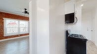 This single-floor space combines classic character with contemporary finishes throughout. The living area serves as the centerpiece of the unit, showcasing a striking full wall of exposed red brick that adds warmth and industrial charm. Multiple windows with white blinds flood the room with natural light, while crown molding, recessed lighting, and a ceiling fan contribute to a polished yet relaxed ambiance. Light wood-tone flooring runs seamlessly throughout the space, unifying each area.

The galley-style kitchen is efficiently designed with crisp white shaker cabinets, dark countertops, and brushed nickel hardware. Black appliances — including a gas range with overhead microwave and a full-size refrigerator — provide a bold contrast to the bright cabinetry. A single-basin stainless steel sink with a modern faucet completes the functional layout.

The bathroom features bold marble-look tile surround at the tub/shower, paired with light wood-tone flooring and a white vanity, creating a clean and elegant feel. A frosted window allows natural light while maintaining privacy.

An additional compact room with white walls and light wood-tone flooring offers flexible use as a bedroom, office, or storage space. Overall, the unit presents a cohesive blend of exposed brick character and updated modern finishes.