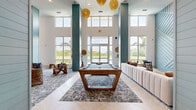 Located in Winter Garden, Florida, this expansive single-floor community clubhouse at Solay Road offers a thoughtfully designed gathering space with a striking coastal-modern aesthetic. The space is defined by soaring double-height ceilings supported by bold teal shiplap columns that serve as dramatic architectural focal points throughout. Large floor-to-ceiling windows flood the interior with natural light while framing lush outdoor views of tropical palms and a tranquil waterway beyond.

Underfoot, light-toned herringbone tile flooring transitions seamlessly into patterned area rugs, adding texture and visual warmth. The open layout encompasses a generous living and lounge area furnished with a large sectional sofa, woven rattan chairs, and curated accent pieces in a coastal palette of cream, blue, and natural wood tones. A mounted flat-screen television anchors one lounge zone, while an upper-level mezzanine balcony overlooks the main floor below.

The space also features a dedicated game area equipped with a statement billiards table and a foosball table, catering to active recreation. Statement gold woven pendant lights hang dramatically from the ceiling, complementing the teal column accents. Intimate nooks with small café-style tables offer quieter retreat spots with serene water views, making this clubhouse a versatile and inviting communal destination.