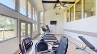 Located at 515 George Wallace Dr in Gadsden, Alabama, this fitness center is designed to provide a bright, energizing environment for exercise. The space is defined by its dramatic vaulted ceilings with exposed beam detailing and large ceiling fans that promote airflow throughout the room. Expansive floor-to-ceiling windows line the walls, flooding the interior with natural light and offering unobstructed views of the surrounding green landscape, creating an open and inviting atmosphere.

The fitness area is well-equipped with a variety of cardio machines, including multiple treadmills and elliptical trainers arranged to maximize use of the space. A wall-mounted flat-screen television is positioned for easy viewing during workouts. For strength training, the space includes a dumbbell rack with a full range of free weights and an adjustable workout bench, providing options for a comprehensive fitness routine.

The flooring throughout is light-toned tile, contributing to the clean and airy aesthetic. A set of French doors leads to an outdoor area, further enhancing the connection to the natural surroundings. Upper-level interior windows hint at an adjoining mezzanine or connected building structure. The overall design balances functionality with an uplifting, well-lit environment suitable for a full range of workout activities.