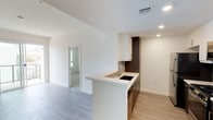 This single-floor residence offers a clean, contemporary design with an open-concept layout that promotes a bright and airy atmosphere. The kitchen is a standout feature, showcasing a peninsula-style breakfast bar with a light-toned countertop, a built-in sink, and a combination of white upper cabinetry and rich dark wood lower cabinets. A mosaic tile backsplash adds a subtle decorative accent, complementing the modern aesthetic. Recessed lighting illuminates the space efficiently, while a ceiling vent ensures comfortable climate control throughout. Light wood-style flooring flows seamlessly across the main living area, adding warmth to the otherwise crisp, white interior. The open living area extends naturally from the kitchen, offering a flexible and spacious feel. A doorway visible from the main space leads to an additional room, hinting at a well-connected and thoughtfully arranged floor plan. The consistent use of neutral tones — from the white walls to the light floors — creates a cohesive, minimalist environment that is both functional and visually appealing, making it an ideal space for modern living.