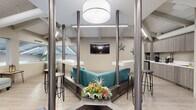 Located in Huntington Beach, California, this coastal-inspired space makes a strong first impression with a striking staircase entry flanked by sleek stainless steel cable railings, leading up into an open, well-appointed interior. The space is defined by dramatic vaulted ceilings with exposed painted beams and tongue-and-groove paneling, lending an airy, spacious feel throughout. Light wood-look flooring runs continuously across the floor, complementing the soft neutral color palette of warm grays and whites.

The main area features a central lounge hub with a teal sectional sofa framed by industrial-style metal columns and topped with a large pendant drum light. Bar-height counters and stools surround the seating area, creating an inviting gathering space. A wall-mounted flat-screen television and coastal starfish décor add personality to the environment. Adjacent to the lounge is a compact kitchenette equipped with built-in appliances and taupe cabinetry.

A separate reception or waiting area includes a cushioned curved chair and a view through interior glass windows overlooking an outdoor pool area, flooding the space with natural light. A treatment or consultation area with a padded bench and dining table with vibrant green seating adds further functionality. The space is finished with linear LED lighting throughout, enhancing its clean, modern, and welcoming atmosphere.