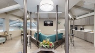 Located in Huntington Beach, California, this coastal-inspired space makes a strong first impression with a striking staircase entry flanked by sleek stainless steel cable railings, leading up into an open, well-appointed interior. The space is defined by dramatic vaulted ceilings with exposed painted beams and tongue-and-groove paneling, lending an airy, spacious feel throughout. Light wood-look flooring runs continuously across the floor, complementing the soft neutral color palette of warm grays and whites.

The main area features a central lounge hub with a teal sectional sofa framed by industrial-style metal columns and topped with a large pendant drum light. Bar-height counters and stools surround the seating area, creating an inviting gathering space. A wall-mounted flat-screen television and coastal starfish décor add personality to the environment. Adjacent to the lounge is a compact kitchenette equipped with built-in appliances and taupe cabinetry.

A separate reception or waiting area includes a cushioned curved chair and a view through interior glass windows overlooking an outdoor pool area, flooding the space with natural light. A treatment or consultation area with a padded bench and dining table with vibrant green seating adds further functionality. The space is finished with linear LED lighting throughout, enhancing its clean, modern, and welcoming atmosphere.