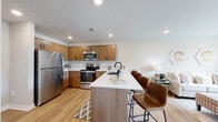 This single-floor residence is designed with a contemporary aesthetic that emphasizes clean lines, warm tones, and functional open-concept living. The kitchen serves as the centerpiece of the main living area, outfitted with flat-front wood cabinetry in a warm walnut finish, crisp white quartz countertops, and a full suite of stainless steel appliances including a refrigerator, range, microwave, and dishwasher. A peninsula island with seating provides an ideal casual dining space while visually connecting the kitchen to the living area. Light hardwood flooring runs throughout the main living spaces, enhancing the bright, airy atmosphere created by recessed lighting and natural light from the windows.

The bedroom is a calm, carpeted retreat featuring a window with an outdoor view, providing natural light. It includes access to a walk-in closet equipped with wire shelving systems for organized storage. The bathroom features a wood-tone vanity with a white countertop, undermount sink, and matte black fixtures, adding a refined modern contrast. A dedicated utility room houses a stacked washer and dryer alongside a water heater and additional wire shelving, offering practical in-unit storage and laundry convenience.