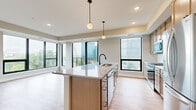 This contemporary single-floor residence is defined by clean lines, abundant natural light, and thoughtful design throughout. The open-concept kitchen serves as the heart of the home, anchored by a large center island with a quartz countertop and an undermount sink. Warm wood cabinetry, a classic subway tile backsplash, and a full suite of stainless steel appliances — including a refrigerator, range, microwave, and dishwasher — complete the space. Globe pendant lights hang above the island, adding a modern, refined touch. Light hardwood flooring flows throughout the main living areas, lending warmth and continuity. Floor-to-ceiling black-framed windows frame striking views of the surrounding urban skyline and lush tree canopy, flooding the interior with natural light. A sliding glass door opens onto a private balcony, extending the living space outdoors. The bedrooms are carpeted and serene, each offering generous closet space, including a spacious walk-in closet with wire shelving and hanging rods. The bathroom features a wood-toned vanity with a white countertop and clean, neutral finishes. Throughout, the neutral color palette and modern fixtures create a polished, move-in-ready atmosphere ideal for urban living.