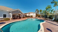 Located at 2209 Minnesota Ave in DeFuniak Springs, Florida, this outdoor amenity area centers around a generously sized freeform swimming pool with a classic kidney-inspired shape and a gradual entry point. The pool is framed by a well-maintained brick paver deck that provides ample space for lounging, with multiple chaise lounge chairs and patio seating arranged throughout the area.

The single-story clubhouse building anchors the space with a welcoming architectural design that features a gabled roofline with warm-toned shingles, decorative circular dormer accents, and large multi-pane windows that allow abundant natural light inside. French doors open directly onto the pool deck, creating a seamless indoor-outdoor connection. A covered patio area adjacent to the building offers shaded seating with umbrella tables, ideal for outdoor dining or relaxing out of the sun.

The pool area is fully enclosed by a black wrought-iron fence, ensuring both safety and security. Elegant lamp posts are positioned around the perimeter, providing lighting for evening use. The surrounding landscape features mature Florida pine and palm trees that create a natural, lush backdrop against the open blue sky, enhancing the overall resort-like ambiance of the space. A grill station is also available on the deck, adding to the outdoor entertainment appeal of this community amenity area.