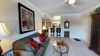 This single-floor residence offers a thoughtfully designed open-concept layout that seamlessly connects the main living area, dining space, and kitchen. The living room is a comfortable and stylish focal point, featuring plush carpeted flooring, crown molding details, and a ceiling fan that enhances the room's airy feel. A large, bold botanical artwork anchors one wall, adding a rich splash of color alongside the deep-toned sofa accented with vibrant red and neutral throw pillows. A round glass coffee table sits at the center, lending a modern touch to the space. The open kitchen is visible from the living area and features white cabinetry, a built-in breakfast bar with tall bar stools, and recessed lighting overhead, making it both functional and inviting. A compact dining nook is tucked adjacent to the kitchen, offering an intimate setting for meals. A decorative mirror and ornate accent cabinet add character to the transitional hallway area, tying together the warm, traditional aesthetic that flows throughout the home. Natural light filters through the space, complementing the neutral wall tones and carpet to create a cohesive, welcoming ambiance throughout.