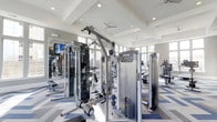 This fitness center offers a comprehensive and thoughtfully designed workout environment for users of all fitness levels. The space features an impressive vaulted ceiling with coffered detailing and recessed lighting, complemented by ceiling fans that ensure comfortable airflow throughout. Large windows line the walls, flooding the interior with natural light and creating an open, energizing atmosphere. The floor is finished with a stylish multi-toned carpet that adds visual interest while providing comfortable footing. The strength training area is anchored by a professional-grade Precor cable crossover machine, alongside a functional trainer and a range of additional strength equipment. Full-length mirrors are mounted along one wall, enhancing the sense of space and allowing users to monitor their form. The cardio section, visible toward the rear of the facility, is lined with multiple machines including treadmills and steppers, all positioned to take advantage of the natural light. The overall layout is spacious and well-organized, allowing for easy movement between equipment. The clean, modern aesthetic and high-quality commercial-grade equipment make this fitness center a standout amenity built to support a wide range of training goals.