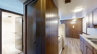 This single-floor unit offers a compact and functional layout distinguished by its warm, wood-paneled walls throughout — a hallmark of its classic design aesthetic. The galley-style kitchen is well-appointed with abundant dark wood cabinetry extending to the ceiling on both sides, a double-basin sink with light-toned countertops, and hardwood-style flooring that adds warmth underfoot. A full-size refrigerator anchors the kitchen, and the layout provides a practical, efficient workflow for everyday use. Adjacent to the kitchen, a hallway connects the main living areas, maintaining the cohesive wood-paneled character of the unit. The bedroom is a generously proportioned space featuring plush carpeting, a textured ceiling, and a dedicated closet area with a rod for hanging storage — providing practical organization. A window in the bedroom frames a pleasant outdoor view of greenery, allowing natural light to filter into the room. The bathroom is clean and straightforward, featuring a combined tub and shower unit, a toilet, a vanity with storage, and light-toned tile flooring. Overall, the unit presents a retro-inspired interior with a consistent design language, offering a functional and livable space well-suited for those seeking a practical, single-level layout.