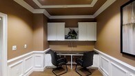 This single-floor space is designed as a functional and polished workspace. The room is finished with warm brown walls accented by crisp white wainscoting panels that line the lower portion of the walls, lending a classic and refined aesthetic. A tray ceiling with white crown molding adds architectural detail and a sense of depth overhead.

The focal point of the room is a built-in workstation featuring a dark granite or stone-topped desk that spans the width of the back wall, providing ample surface area for productivity. Two black leather armchairs with sled bases are positioned at the desk, accommodating collaborative or dual-user work. Above the desk, a series of white upper cabinets flank a central open display niche, which features decorative greenery, offering both practical storage and visual interest.

The space is finished with neutral carpet flooring that complements the overall color palette. Recessed lighting in the tray ceiling ensures even illumination throughout the room. The overall ambiance is professional and organized, making this workspace well-suited for focused work or meetings in a compact, efficiently designed environment.
