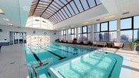This elevated aquatic facility is defined by its striking architectural design and sweeping urban vistas. At its centerpiece is a full-length lap pool with clearly marked lanes, accompanied by a separate hot tub/wading area with entry steps. A dramatic barrel-vaulted skylight spans the length of the pool ceiling, flooding the space with natural light and enhancing the open, airy atmosphere.

The pool deck is finished in large-format light-colored tiles and features lounge chairs, small seating arrangements, and potted greenery positioned along the perimeter. A prominent stone-clad accent wall adds a warm, textural contrast to the otherwise crisp white interior. Large floor-to-ceiling windows line the facility on multiple sides, offering unobstructed panoramic views of a major city skyline. An access point leads to what appears to be an outdoor terrace area.

Adjacent to the pool is a dedicated lounge zone featuring modern white upholstered sofas and chairs arranged around accent tables, positioned directly in front of expansive floor-to-ceiling windows that frame dramatic skyline views. The light tile flooring, white structural columns, and abundant natural light throughout both areas contribute to a bright, sophisticated, and resort-like ambiance.
