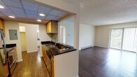 Located at 799 W Fifth Ave in Naperville, IL, this single-floor residence offers a well-organized layout designed for comfortable everyday living. The kitchen is a functional galley-style space featuring warm wood cabinetry, dark granite countertops with a mosaic tile backsplash, stainless steel appliances including a refrigerator and range, and a drop-panel ceiling with recessed lighting. A pass-through opening connects the kitchen to the main living area, enhancing the open feel between spaces. The living room is generously sized, featuring rich dark hardwood-style flooring, a textured ceiling, a ceiling fan, and a baseboard heating system. A sliding glass door floods the space with natural light and provides direct access to an outdoor balcony with views of surrounding greenery. The bedrooms are carpeted and offer a cozy, quiet atmosphere, each equipped with a ceiling fan and vertical blinds over windows that overlook outdoor greenery. Closet space is provided within the bedrooms, including a walk-in style closet with a rod and open shelving. The bathroom is clean and straightforward, featuring a full soaking tub with an overhead shower, white square tile surround, and chrome fixtures. Throughout the unit, warm beige walls and consistent finishes create a cohesive and inviting ambiance.