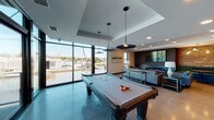 This contemporary single-floor amenity space is thoughtfully designed to offer a premium social and recreational experience, highlighted by sweeping waterfront views of a nearby marina. Floor-to-ceiling windows in the billiards area flood the space with natural light while framing picturesque views of the water and docked boats, creating a bright and inviting atmosphere. A full-size pool table anchors this zone, complemented by stylish pendant lighting overhead. Adjacent to the billiards area is a comfortable lounge seating area furnished with rich brown leather sofas and armchairs, with large windows continuing the marina views. The main lounge features a sophisticated layout with grey upholstered seating, glass coffee tables, a sleek wall-mounted fireplace, a large flat-screen TV set against dark wood paneling, and a cozy booth dining nook framed by warm wood-accent walls and globe pendant lighting. The kitchen area is well-appointed with a large dark-topped island with bar seating, white subway tile backsplash, open metal shelving, warm wood cabinetry, stainless steel appliances including a full-size refrigerator, and an additional wall-mounted display screen — ideal for catered events or casual gatherings. The overall design blends modern industrial elements with warm finishes throughout.