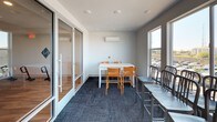 This dedicated study room offers a clean, functional environment designed to support productive work and collaboration. The space features a contemporary aesthetic with crisp white walls, recessed lighting, and dark, textured carpet tiles that add a grounded, professional feel underfoot. A wall-mounted mini-split air conditioning unit ensures year-round climate comfort, making the room suitable for extended study sessions regardless of the season.

The room is furnished with a white rectangular table accompanied by vibrant orange chairs, providing a bright and energizing focal point. Additional seating is available with a set of gray metal chairs arranged along a separate table area near the windows, allowing the space to accommodate varying group sizes and configurations.

Large, floor-to-ceiling windows along one wall flood the room with abundant natural light and frame views of the surrounding neighborhood and skyline beyond. A full-height glass partition wall with a sliding glass door separates the study room from an adjacent corridor or common area, maintaining visual openness while offering acoustic separation for focused work. Wall-mounted electrical outlets are conveniently positioned to support device charging and connectivity needs. Overall, the study room strikes a balance between functional simplicity and an inviting, modern atmosphere.