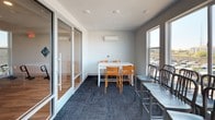 This dedicated study room offers a clean, functional environment designed to support productive work and collaboration. The space features a contemporary aesthetic with crisp white walls, recessed lighting, and dark, textured carpet tiles that add a grounded, professional feel underfoot. A wall-mounted mini-split air conditioning unit ensures year-round climate comfort, making the room suitable for extended study sessions regardless of the season.

The room is furnished with a white rectangular table accompanied by vibrant orange chairs, providing a bright and energizing focal point. Additional seating is available with a set of gray metal chairs arranged along a separate table area near the windows, allowing the space to accommodate varying group sizes and configurations.

Large, floor-to-ceiling windows along one wall flood the room with abundant natural light and frame views of the surrounding neighborhood and skyline beyond. A full-height glass partition wall with a sliding glass door separates the study room from an adjacent corridor or common area, maintaining visual openness while offering acoustic separation for focused work. Wall-mounted electrical outlets are conveniently positioned to support device charging and connectivity needs. Overall, the study room strikes a balance between functional simplicity and an inviting, modern atmosphere.