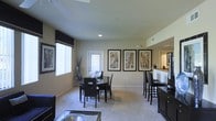 This single-floor residence showcases an open-concept layout that seamlessly connects the living, dining, and kitchen areas into one cohesive space. The dining area is anchored by a round dark-wood table paired with matching black chairs, positioned beneath recessed ceiling lighting that provides warm, focused illumination. Neutral-toned walls are adorned with a curated set of large framed architectural prints, adding an urban, sophisticated character to the space. Plush wall-to-wall carpeting runs throughout the main living areas, contributing to a comfortable and refined atmosphere. Natural light filters in through generously sized windows dressed with fabric valances, brightening the interior. Adjacent to the dining area, a breakfast bar with dark cabinetry and raised seating connects to the kitchen, offering a functional transition between spaces. The kitchen area, visible beyond the bar, features warm-toned countertops and under-cabinet lighting. A ceiling fan overhead ensures year-round comfort. The overall aesthetic is contemporary and cohesive, blending dark furnishings against light walls and flooring for a balanced, inviting ambiance.