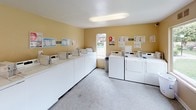 Located at 801 S Lyon St in Santa Ana, California, this single-floor community laundry facility is designed to serve residents with a practical and well-equipped space. The room features warm beige walls, a light-toned speckled floor, and flush overhead fluorescent lighting that keeps the area bright and functional throughout the day.

The facility is lined with a generous bank of front-loading washing machines along one wall, complemented by a set of stacked dryers on the opposite side, providing ample laundry capacity for community use. Each machine is clearly numbered and equipped with coin-operated controls for straightforward use. Instructional and informational signage is posted on the walls for resident reference.

A Coca-Cola vending machine adds a convenient amenity near the dryer bank. A sturdy white folding table is available for sorting and folding laundry, and a wall-mounted fan provides additional air circulation. A fire extinguisher is visibly mounted near the entrance, reflecting attention to safety.

Large windows along one wall allow natural light to fill the space and offer views of the surrounding landscaped grounds, including green lawns and the neighboring building exterior. A single door provides direct access to the outdoor walkway. The overall layout is clean, spacious, and efficiently organized to accommodate the everyday laundry needs of the community.