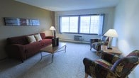 This single-floor space centers around a well-appointed living room that offers a comfortable and functional atmosphere. The room is finished with wall-to-wall carpeting in a neutral tone, providing a soft, understated foundation that complements the overall interior palette. Two-toned walls — warm beige paired with a deeper taupe accent — add visual depth without overwhelming the space.

Large, multi-pane windows along the exterior wall allow abundant natural light to fill the room, creating a bright and open feel. Sheer curtains frame the windows, softening the incoming light while maintaining a sense of airiness. A wall-mounted HVAC unit and baseboard heating elements beneath the windows provide year-round climate control.

The living area is furnished with a deep red sofa accented by neutral throw pillows, a pair of patterned upholstered armchairs, and a rectangular wood-and-metal coffee table that anchors the seating arrangement. A tall decorative branch arrangement adds a natural, organic accent near the window. A floor lamp provides additional ambient lighting. The space is thoughtfully arranged to encourage conversation and relaxation, presenting a balanced and inviting environment overall.