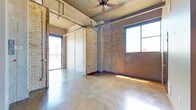 This single-floor unit showcases a compelling industrial aesthetic rooted in its original architectural character. The main living area is defined by polished concrete floors that reflect natural light streaming in through large, metal-framed windows. Exposed brick walls in warm yellow and red tones add texture and depth throughout, while concrete structural columns and fully exposed ductwork, conduit, and ceiling joists overhead reinforce the loft-like industrial atmosphere. A ceiling fan circulates air through the generous open space. A large glass-panel sliding partition offers flexible room division while maintaining the open feel. The kitchen is well-appointed with rich, dark wood cabinetry, a mosaic tile backsplash, marble-style countertops, and a full suite of stainless steel appliances including a refrigerator, electric range, dishwasher, and microwave. A dramatic cylindrical column anchors the kitchen area, adding a striking architectural focal point. The bathroom features a walk-in shower, a dark wood vanity with a marble-top sink, and a wall mirror. Natural light reaches multiple areas of the unit, enhancing the open, airy quality of the space throughout.