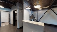 This single-floor space showcases a compelling blend of industrial character and modern finishes. The open main area is defined by its dramatic dark wood plank ceiling with exposed structural steel trusses, which lend a raw, loft-like aesthetic throughout. Polished concrete floors run continuously, reinforcing the industrial ambiance while remaining easy to maintain.

The kitchen is thoughtfully appointed with white shaker-style cabinetry, light quartz countertops, and a vertically stacked blue-gray tile backsplash. Stainless steel appliances — including a freestanding range with a smooth cooktop and a full-size refrigerator — complete the space. A peninsula with a quartz countertop overhang extends into the open area, providing a natural division between the kitchen and the main floor, and is accented by two pendant lights with industrial-style metal shades that hang from the wood ceiling above.

The open main room offers generous, flexible floor space with exposed steel truss framing along one wall and a window that admits natural light and outdoor views, adding warmth to the otherwise industrial setting.

The bathroom features wood-look tile flooring, a white vanity with a quartz countertop and undermount sink, and clean, neutral wall finishes. The space also includes a corridor with closet access, connecting the functional areas efficiently. Overall, this space offers a distinctive industrial character with quality modern upgrades throughout.