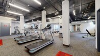 Located at 929 Jefferson St in Kansas City, Missouri, this fitness facility offers a comprehensive workout environment spread across a single, generously sized level. The space is divided into two distinct gym areas, each designed to support a variety of exercise needs.

The main gym area showcases an industrial-inspired aesthetic, with exposed ceilings revealing ductwork and structural beams finished in dark tones, contrasted by bright white columns and walls. Large floor-to-ceiling glass partitions divide sections of the space, maintaining an open, connected feel while allowing natural light to filter through. The flooring throughout consists of durable, patterned carpet tiles. This area is equipped with a row of treadmills, elliptical machines, cable and strength training machines, adjustable benches, and full-length mirrors that enhance the sense of space. Exposed brick accent walls add a warm, urban character to the environment.

The second gym area serves as a dedicated cycling studio, featuring a drop ceiling with recessed lighting for a more intimate atmosphere. A wall-mounted display screen anchors the front of the room, while rows of professional spinning bikes face it directly. Mirrored walls and natural light from windows complete the studio setting.

Together, these two spaces provide a well-rounded fitness facility suited for cardio training, strength work, and group cycling classes.