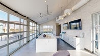 This contemporary single-story amenity space showcases a thoughtfully designed open-concept layout with striking architectural details throughout. Soaring vaulted ceilings create an airy, expansive atmosphere, complemented by exposed ductwork that adds an industrial edge to the modern aesthetic. Light-toned stone tile flooring and white mosaic brick accent walls establish a clean, cohesive visual identity throughout the space.

The kitchen area serves as a central hub, anchored by a large white quartz island with an undermount double sink and stylish pendant lighting overhead. White shaker cabinetry lines the adjacent wall, accompanied by built-in appliances and counter space, while wall-mounted flat-screen monitors add functionality. Full-height glass garage doors open directly to an outdoor pool and lounge deck, flooding the interior with natural light and enabling seamless indoor-outdoor flow.

A lounge area adjacent to the kitchen features comfortable seating arrangements and large glass French doors that further connect the interior to the outdoor environment. A dedicated corridor leads to additional rooms or private spaces. Entry vestibules on multiple sides provide convenient access with generous glazing that maintains the bright, welcoming character of the overall space.