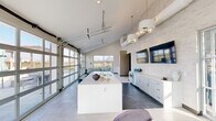 This contemporary single-story amenity space showcases a thoughtfully designed open-concept layout with striking architectural details throughout. Soaring vaulted ceilings create an airy, expansive atmosphere, complemented by exposed ductwork that adds an industrial edge to the modern aesthetic. Light-toned stone tile flooring and white mosaic brick accent walls establish a clean, cohesive visual identity throughout the space.

The kitchen area serves as a central hub, anchored by a large white quartz island with an undermount double sink and stylish pendant lighting overhead. White shaker cabinetry lines the adjacent wall, accompanied by built-in appliances and counter space, while wall-mounted flat-screen monitors add functionality. Full-height glass garage doors open directly to an outdoor pool and lounge deck, flooding the interior with natural light and enabling seamless indoor-outdoor flow.

A lounge area adjacent to the kitchen features comfortable seating arrangements and large glass French doors that further connect the interior to the outdoor environment. A dedicated corridor leads to additional rooms or private spaces. Entry vestibules on multiple sides provide convenient access with generous glazing that maintains the bright, welcoming character of the overall space.