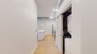This single-floor unit showcases a modern, industrial-influenced design highlighted by exposed ductwork running along the ceiling, which contrasts with the clean white walls and light hardwood flooring throughout the main living areas. The open-concept layout seamlessly connects the kitchen and living space, creating a generous, airy feel with natural light filtering in through windows along the far wall.

The kitchen is well-appointed with white cabinetry featuring matte black hardware, a white subway tile backsplash, stainless steel appliances including a dishwasher, range, and built-in microwave, and a deep utility sink. A large white kitchen island with pendant lighting serves as a central gathering point and provides additional storage and counter space.

A dedicated laundry closet is conveniently positioned just off the kitchen, housing a stacked washer and dryer unit behind sliding doors. The full bathroom is finished in a clean, contemporary style with large-format white floor tiles, a soaking tub and shower combination with floor-to-ceiling beige tile surround, white cabinetry, and matte black fixtures and hardware. An additional carpeted room provides flexible space suitable for use as a bedroom or office. Recessed lighting and pendant fixtures provide consistent illumination throughout.