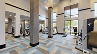 Located at 910 Deerfield Crossing Dr in Alpharetta, GA, this well-appointed fitness center offers a generously sized, single-floor exercise space designed to accommodate a full range of workout routines. The room is immediately distinguished by its soaring double-height ceilings, which lend an open, airy atmosphere throughout. Large floor-to-ceiling windows along the perimeter flood the space with natural light and provide views of the surrounding greenery, creating an energizing environment for exercise.

The flooring features a striking herringbone-pattern carpet tile in a palette of blue, gray, and tan, adding a modern aesthetic to the functional space. Textured column wrappings in a woven metallic finish serve as architectural focal points, complementing the clean white walls accented with dark trim details.

Equipment is thoughtfully arranged across distinct zones. The cardio area houses a row of treadmills, stationary bikes, stair climbers, and elliptical machines, many positioned to face the outdoor views. A dedicated strength training section offers free weights, dumbbells, barbells, adjustable benches, and a cable machine system. Full-length framed mirrors line one wall, enhancing the sense of space and supporting proper form during workouts. A wall-mounted television and ceiling fans with pendant lighting complete the well-designed, functional environment.