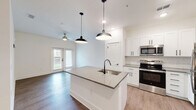 Located at Harbor Pointe Circle in Fruitland Park, FL, this well-designed one-bedroom, one-bathroom unit offers a thoughtful layout across a single floor. The open-concept living area seamlessly connects the kitchen, dining, and living spaces, creating a bright and functional hub for daily life. The kitchen is a standout feature, showcasing white shaker cabinets, subway tile backsplash, stone countertops, stainless steel appliances, and a central island with a matte black sink and pendant lighting overhead. A ceiling fan enhances the living area, which leads to a private patio through glass-panel doors, allowing natural light to flood the space. Wood-look flooring runs throughout the main living areas, while the spacious bedroom features plush carpet, a ceiling fan, a large window with outdoor views, and access to two closets. The full bathroom is finished with a white vanity, stone countertop, and a tub-shower combination. A dedicated laundry room with washer and dryer hookups, a pantry, and a utility room provide excellent storage and convenience. The clean, neutral palette and modern hardware throughout lend a fresh, contemporary feel to this well-appointed home.