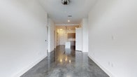 This single-floor space showcases a clean, modern aesthetic defined by its seamless open layout and striking polished concrete floors that reflect light throughout the interior. The expansive main living area flows effortlessly into the kitchen, creating an open, connected feel ideal for everyday living and entertaining.

The kitchen is a standout feature, equipped with dark wood cabinetry that provides a warm contrast against the bright white walls and surfaces. A central island anchors the space, complemented by stylish pendant lighting that adds a contemporary touch. Recessed lighting throughout ensures the space remains bright and well-illuminated.

The crisp white walls and clean lines of the interior contribute to a minimalist, modern ambiance, while the polished concrete flooring adds an industrial edge that ties the design together cohesively. Visible HVAC vents and thoughtful ceiling fixtures reflect practical yet stylish design choices throughout.

Wide doorways and an open corridor enhance the sense of spaciousness, making the layout feel airy and unobstructed. Overall, this space combines industrial and contemporary design elements to create a functional, visually compelling environment with a refined, urban character.