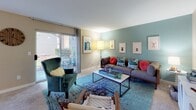 This single-floor residence offers a thoughtfully designed layout that balances comfort and functionality across its key living areas. The living room serves as the heart of the home, featuring a soothing teal accent wall, soft carpeted flooring, and a sliding glass door that opens to an outdoor patio, allowing natural light to fill the space. The room provides ample space for seating and is enhanced by tasteful wall art and warm ambient lighting. Adjacent to the living area, the galley-style kitchen is equipped with white cabinetry featuring wood trim, granite-style countertops, a black electric range, stainless steel refrigerator, and wood-look vinyl flooring — offering a clean and efficient cooking environment with dedicated counter space on both sides. The bedroom is a calm retreat with plush carpet, neutral beige walls, and a window dressed with decorative patterned curtains that frame an outdoor view, complemented by framed artwork adding a personal touch. The full bathroom is well-appointed with wood-look vinyl flooring, a combined tub and shower with a decorative curtain, a vanity with granite-style countertops, and a large mirror that enhances the sense of space. Together, these spaces create a cohesive, comfortable living environment with a modern aesthetic.