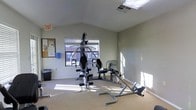 This single-floor fitness room offers a functional workout environment designed to accommodate a range of exercise needs. The space features vaulted ceilings that lend an open, airy feel, complemented by natural light streaming in through a window that provides a view of the surrounding exterior. The light-toned walls and carpeted flooring create a clean, neutral backdrop throughout the room.

The equipment layout is well-organized, centering around a multi-station cable and weight machine suitable for a variety of strength training exercises. Alongside it, a rowing machine and an abdominal/core trainer provide options for cardiovascular and targeted muscle conditioning. A bulletin board mounted near the entrance displays informational materials, adding a practical touch to the space. An emergency exit sign is clearly posted, and a ventilation grille in the ceiling ensures proper airflow during workouts.

A waste bin and a wall-mounted dispenser — likely for sanitizing wipes or paper towels — reflect attention to basic hygiene and maintenance standards. The room is straightforward in its purpose, offering a no-frills but adequately equipped setting for residents or users looking to maintain an active lifestyle without leaving the property.