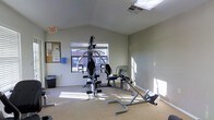 This single-floor fitness room offers a functional workout environment designed to accommodate a range of exercise needs. The space features vaulted ceilings that lend an open, airy feel, complemented by natural light streaming in through a window that provides a view of the surrounding exterior. The light-toned walls and carpeted flooring create a clean, neutral backdrop throughout the room.

The equipment layout is well-organized, centering around a multi-station cable and weight machine suitable for a variety of strength training exercises. Alongside it, a rowing machine and an abdominal/core trainer provide options for cardiovascular and targeted muscle conditioning. A bulletin board mounted near the entrance displays informational materials, adding a practical touch to the space. An emergency exit sign is clearly posted, and a ventilation grille in the ceiling ensures proper airflow during workouts.

A waste bin and a wall-mounted dispenser — likely for sanitizing wipes or paper towels — reflect attention to basic hygiene and maintenance standards. The room is straightforward in its purpose, offering a no-frills but adequately equipped setting for residents or users looking to maintain an active lifestyle without leaving the property.