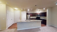 This single-floor unit is thoughtfully designed with an open-concept layout that seamlessly connects the kitchen and living areas. The kitchen serves as a central highlight, featuring rich dark wood cabinetry, granite countertops, and stainless steel appliances including a refrigerator and over-the-range microwave. A black electric range adds contrast, while a generously sized granite-topped island with an integrated sink provides additional workspace and functions as a natural divider between the kitchen and living space. Track lighting illuminates the kitchen and dining area effectively.

The living area is carpeted and benefits from natural light through a set of glass doors leading to an outdoor access point, complemented by a ceiling fan overhead. The bedrooms are similarly carpeted, each featuring a ceiling fan and a window with blinds that allow natural light to filter in.

The primary bathroom offers a dual-sink vanity with dark cabinetry, a large mirror with strip lighting above, and a separate shower and tub combination. A secondary bathroom provides a single-sink vanity with matching dark cabinetry, tile flooring, and a well-lit mirror. Throughout the unit, neutral wall tones and consistent dark cabinetry create a cohesive, functional aesthetic.