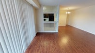 This single-floor residence offers a thoughtfully designed open layout that seamlessly connects the living, dining, and kitchen areas. Rich, warm hardwood-style flooring runs throughout the main living spaces, lending a cohesive and inviting feel to the entire unit.

The living area is bathed in natural light through large windows dressed with vertical blinds, providing both brightness and privacy. A distinctive white spindle partition wall separates the living space from the kitchen, maintaining an open and airy connection between rooms while adding a charming architectural detail.

The kitchen features dark cabinetry paired with a light countertop, and is equipped with a full-size refrigerator and microwave. The raised bar-style counter provides a casual dining or prep surface and reinforces the open-concept flow.

Adjacent to the kitchen, the dining area is elevated in elegance by a classic multi-arm chandelier that serves as a focal point. Wainscoting along the dining room walls adds a refined, traditional touch to the overall design.

Throughout the space, the combination of warm flooring tones, white trim work, and natural light creates a welcoming and functional environment suited for everyday living and entertaining alike.