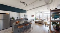 This contemporary single-floor residence showcases an open-concept layout that seamlessly connects the kitchen, dining, and living areas. The kitchen is a standout feature, equipped with stainless steel appliances, two-tone cabinetry in white and gray, a subway tile backsplash, a bold teal accent wall, and a large island with a dark stone countertop and wooden bar stools — ideal for both cooking and casual entertaining. Exposed industrial-style ceiling ducts and track lighting add an urban loft character throughout.

The living area is bathed in natural light from expansive floor-to-ceiling black-framed windows, offering sweeping elevated city views. A balcony door extends the living space outdoors. Light wood-tone flooring runs throughout, enhancing the airy, cohesive feel.

The bedroom is a serene retreat, featuring large windows with the same striking cityscape panorama, high ceilings, and clean white walls. The bathroom is thoughtfully finished with hexagonal tile flooring, white subway tile walls, a gray vanity with matte black fixtures, and a framed mirror. A stacked in-unit washer and dryer provide added convenience. Overall, the space delivers a polished, modern aesthetic with industrial touches and abundant natural light.