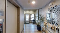 This single-floor space presents a polished and professional environment designed with both function and comfort in mind. The entryway sets a welcoming tone, featuring light hardwood-style flooring that extends throughout, neutral-toned walls, and recessed ceiling lighting that provides consistent, even illumination. A glass-paneled entry door allows natural light to flow into the corridor, brightening the interior and creating an open, airy atmosphere. The hallway is well-appointed, with a sleek black accent chair offering a seating area near the entrance, ideal for receiving visitors. A large potted palm plant adds a touch of greenery and warmth to the space, softening the clean lines of the modern interior. Security features are integrated discreetly into the ceiling, including a dome camera and sprinkler system, reflecting a commitment to safety without disrupting the aesthetic. Display materials and informational signage are neatly arranged along one wall, suggesting a client-facing or public-service orientation. The overall ambiance is contemporary and well-maintained, combining practical design elements with inviting touches that make the space feel both approachable and efficient.