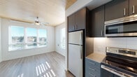 Located at 1090 NE 91st Ave in Hillsboro, Oregon, this thoughtfully designed single-floor unit showcases a contemporary aesthetic with warm, natural wood-paneled ceilings that serve as a defining architectural feature across the living spaces. Light-toned luxury vinyl plank flooring flows throughout the main living areas, complemented by crisp white walls that enhance the bright, airy atmosphere created by multiple windows with horizontal blinds allowing generous natural light.

The kitchen is a standout feature, offering two distinct configurations visible throughout the space. One version presents dark charcoal cabinetry paired with a stainless steel refrigerator and microwave, while another showcases deep navy-toned cabinetry with a full suite of stainless steel appliances including a range, dishwasher, and refrigerator. Both kitchens feature clean white subway tile backsplashes and ample upper and lower cabinet storage.

The bedroom areas include carpet flooring and ceiling fans, with windows overlooking outdoor greenery. The bathroom is well-appointed with a modern dark-toned vanity, quartz-style countertop, a large mirror with contemporary strip lighting, and direct access to a walk-in closet with built-in shelving and hanging rods. Recessed lighting and ceiling fans throughout add to the functional, modern character of this well-designed Hillsboro residence.