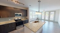 This single-floor residence on Brickworks Drive in Cayce, South Carolina, presents a contemporary open-concept design that seamlessly integrates the kitchen, dining, and living areas. The kitchen serves as a central highlight, featuring a large granite-topped island with an undermount sink and pendant lighting overhead. Dark-toned flat-front cabinetry extends to the ceiling, providing generous storage, while a glass tile backsplash and stainless steel appliances — including a range, microwave, and refrigerator — complete the modern aesthetic. Light wood-look flooring flows throughout the main living areas, enhancing the bright and airy atmosphere created by multiple windows fitted with horizontal blinds. The living area is generously sized, anchored by a ceiling fan and French doors that open to a private balcony, offering outdoor views. The bedroom features plush carpeting, a ceiling fan, and a window that brings in natural light. The bathroom is well-appointed with a granite-topped dark wood vanity, a large framed mirror with overhead lighting, and a tub-shower combination. A dedicated laundry area houses a full-sized washer and dryer with overhead shelving, adding practical convenience to this thoughtfully designed space.