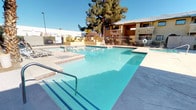 This outdoor amenity complex offers a comprehensive set of recreational facilities designed for active and leisurely enjoyment under clear desert skies. At the heart of the space is a generously sized swimming pool with vivid turquoise water, complemented by a dedicated spa and hot tub area that features active water jets — ideal for relaxation. The pool deck is finished in smooth concrete and is equipped with lounge chairs, providing ample space for sunbathing and socializing. Entry to the pool is made convenient with built-in steps and stainless steel handrails.

The area is enclosed by a secure metal perimeter fence, ensuring privacy and safety throughout. Mature palm trees, pines, and desert-adapted shrubs are thoughtfully landscaped around the space, offering both shade and a lush aesthetic contrast to the arid surroundings. A utility or storage structure sits nearby, adding practical functionality to the complex.

Adjacent to the pool area is a full-sized tennis court, surfaced in a classic green and red color scheme with clearly marked lines and a regulation net. The court is outfitted with overhead lighting for evening play and is framed by a chain-link enclosure. A distinctive white tower structure serves as a charming architectural focal point near the court, enhancing the overall character of the facility.