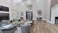 This elegantly designed single-floor communal space blends classic architectural details with contemporary styling to create a refined and welcoming environment. The centerpiece of the main lounge is its dramatic double-height ceiling, adorned with a crystal chandelier that casts a warm glow over the expansive open floor plan. Smooth white walls, crisp crown molding, and wide-plank wood-look flooring establish a clean, sophisticated aesthetic throughout.

The main seating area is thoughtfully arranged with gray tufted and swivel lounge chairs, a geometric-patterned area rug, and a wood-and-metal coffee table, all anchored by a striking cobalt blue accent wall featuring a grand arched window. This statement window floods the space with natural light and frames outdoor views of lush greenery. Framed artwork is displayed throughout, adding cultural character to the walls.

An exposed brick accent wall transitions naturally into a bar-height work counter with barstool seating, ideal for collaborative use. The adjacent kitchen area is well-appointed with dark cabinetry featuring decorative cross detailing, quartz countertops, a stainless steel farmhouse sink, dishwasher, and a beverage station. A large white communal table with side seating provides ample workspace or dining use. Black-framed glass entry doors complete the polished, upscale character of this versatile space.