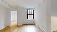 This thoughtfully designed single-floor residence presents a clean, contemporary aesthetic defined by crisp white walls and light-toned hardwood flooring that flows seamlessly through the living areas and bedrooms. The open layout connects a well-proportioned living space to a compact, fully equipped kitchen featuring white cabinetry with matte black hardware, granite countertops, classic subway tile backsplash, and a full suite of stainless steel appliances including a range, microwave, and dishwasher. Natural light enters through black-framed windows that offer views of the surrounding brick building facades, lending an urban character to the interior. The bedrooms are well-sized, with clean lines and recessed linear lighting adding a modern touch to the ceilings. The bathroom is finished with large-format floor tiles, white subway tile walls, a sleek rectangular sink mounted on a dark wood vanity, and a wall-mounted mirror with accent lighting. The unit is accessed through a striking dark-paneled entry door, and the overall palette of whites, light wood, and black accents creates a cohesive, modern sensibility throughout. The space is move-in ready and designed for comfortable, functional urban living.