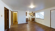 This single-story space features an open-plan layout that seamlessly connects the living area to the kitchen, creating a functional and cohesive environment. The flooring throughout is a rich, dark hardwood-style material that adds warmth and continuity across the space.

The kitchen is positioned along the back wall and is equipped with medium-toned wood cabinetry, both upper and lower, providing ample storage. White appliances, including a refrigerator and electric range, are neatly integrated beneath a range hood. A peninsula or partial counter division subtly separates the kitchen from the living area while maintaining the open feel.

A large window on the right-hand wall admits generous natural light, brightening the entire space. White walls and white baseboards provide a clean, neutral backdrop throughout. A doorway on the left leads to an additional room, partially visible as a hallway or closet space with matching flooring and trim, suggesting further utility areas beyond.

The ceiling features a flush-mount light fixture and a textured finish. Overall, the space presents a straightforward, well-organized layout with a warm, neutral aesthetic suited to everyday living.