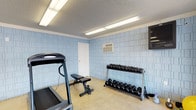 This single-story fitness room offers a functional workout environment designed to accommodate a range of exercise routines. The walls are finished with light blue tile block, giving the space a clean, utilitarian appearance. Flooring consists of a neutral-toned carpet, providing a modest cushioned surface throughout the room. Overhead, fluorescent strip lighting ensures consistent, bright illumination across the entire area. A wall-mounted air conditioning unit provides climate control, keeping the space comfortable during workouts. The room is equipped with a treadmill positioned near one wall, an adjustable weight bench centrally placed for versatile strength training, and a full multi-tier dumbbell rack stocked with a range of free weights. A wall-mounted storage unit on one side offers additional organizational space. A single door provides access to and from the room. The textured ceiling and efficient layout reflect a no-frills, practical approach to fitness facility design, making this space well-suited for straightforward cardio and strength training sessions.