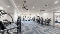 Located at 1533 Ellis Rd in Durham, North Carolina, this fitness center offers a generously sized, single-floor exercise room designed to support a well-rounded workout routine. The space is outfitted with a wide variety of equipment, including multiple treadmills, elliptical machines, a stationary bike, cable weight machines, selectorized strength stations, and a dedicated free weights area complete with a dumbbell rack and adjustable bench.

The interior design is clean and modern, featuring crisp white walls accented by subtle gray crown molding and a distinctive shiplap wall panel that adds a touch of texture. The floor is covered in stylish, patterned carpet tiles in cool tones of blue, gray, and teal, lending the space a polished, contemporary feel. Recessed lighting is distributed throughout the ceiling, supplemented by multiple ceiling fans that help maintain a comfortable environment during workouts.

Natural light filters in through several windows along the perimeter, offering outdoor views and a bright, airy ambiance. Interior windows provide additional visual connectivity to adjacent areas of the facility. A glass-paneled door provides convenient access to the exterior. The overall atmosphere is welcoming and functional, making it a well-equipped destination for residents seeking a complete fitness experience.